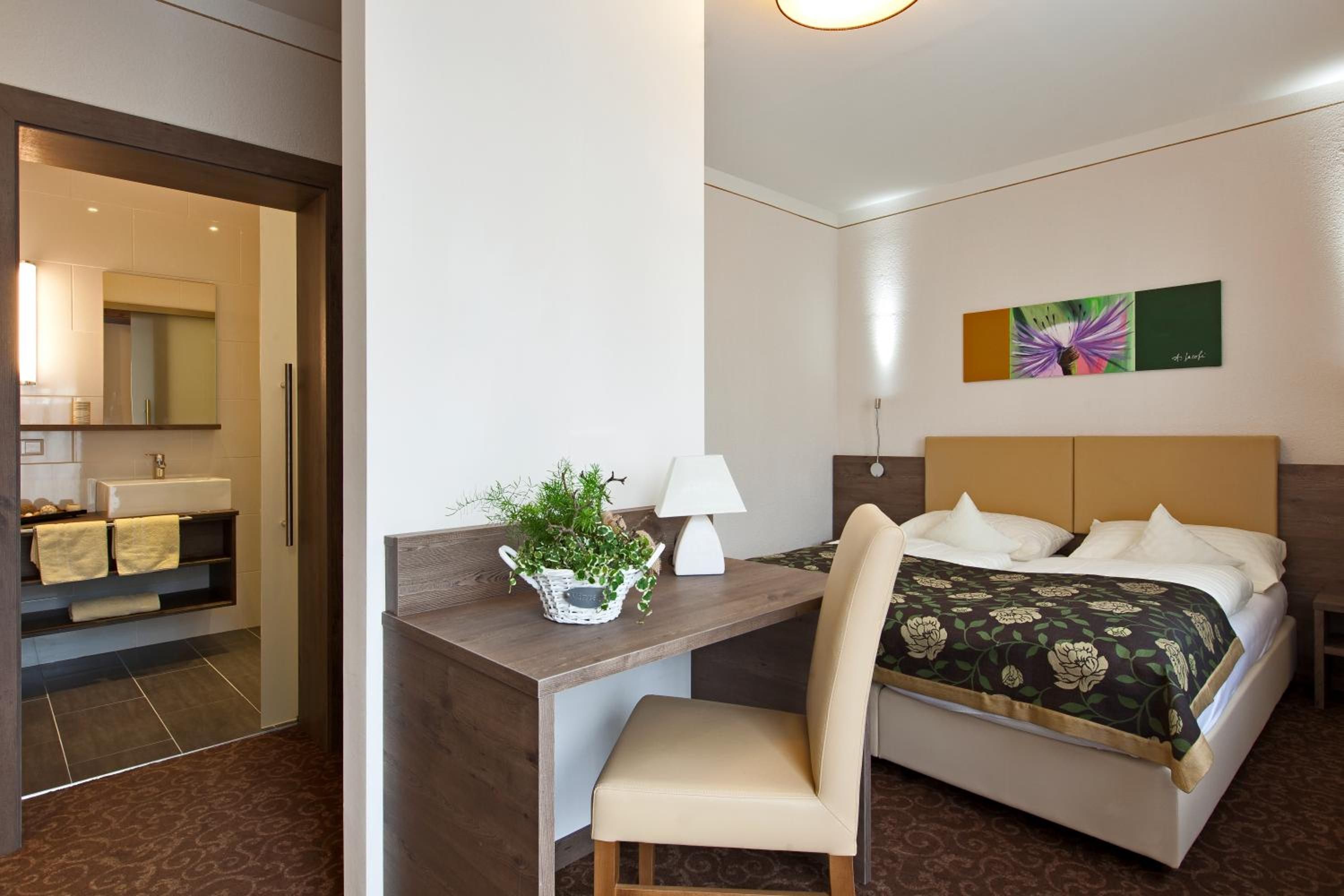 Ein Hotelzimmer mit einem Bett, einem Schreibtisch mit Stuhl, einer Lampe, einer Pflanze und einem Bild an der Wand.