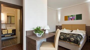 Ein Hotelzimmer mit einem Bett, einem Schreibtisch mit Stuhl, einer Lampe, einer Pflanze und einem Bild an der Wand.