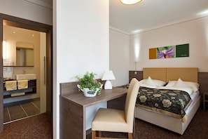 A hotel room with a bed, a desk with a chair, a lamp, a plant, and a painting on the wall.
