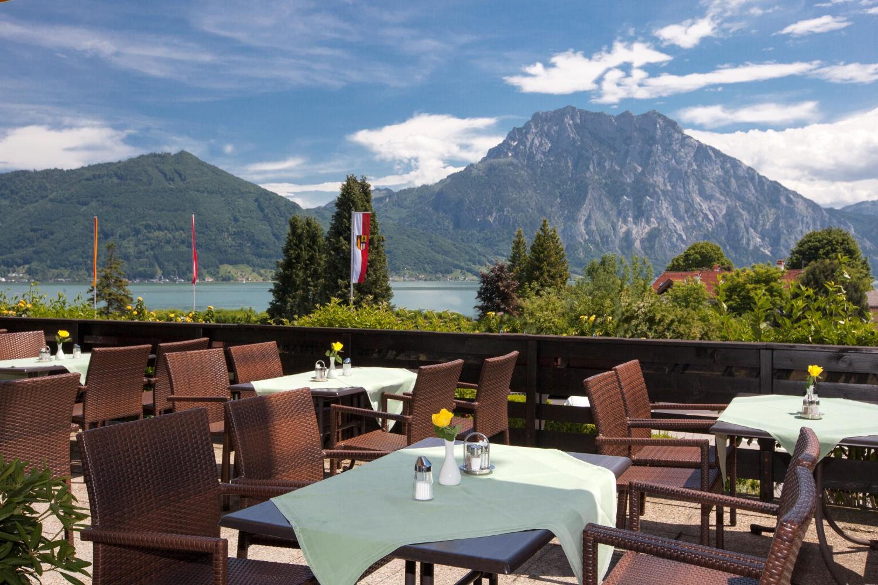 An outdoor dining area with wicker chairs and tables, overlooking a lake and mountains.