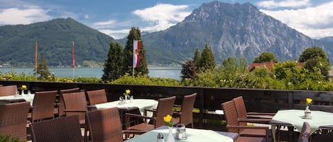 An outdoor dining area with wicker chairs and tables, overlooking a lake and mountains.