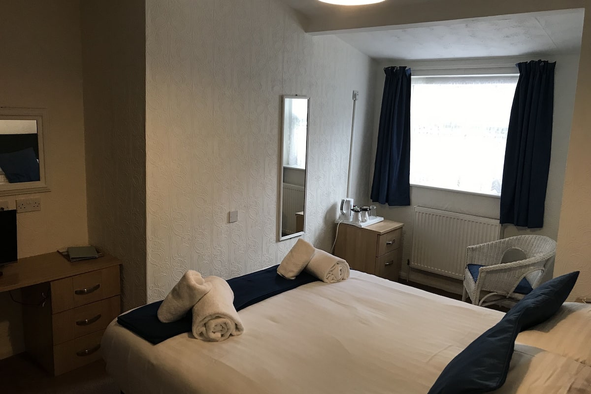 Quarto de hotel com cama, mesa de cabeceira, espelho, janela com cortinas e uma cadeira.