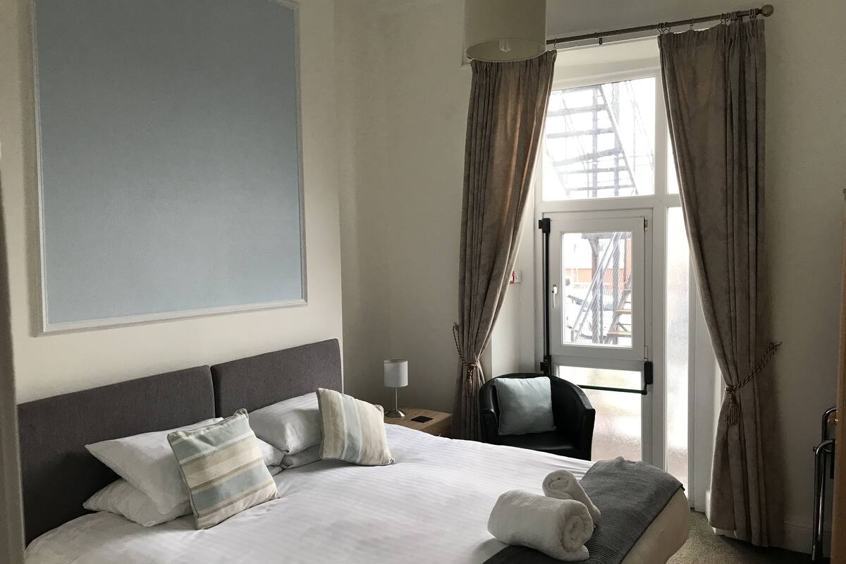 Quarto de hotel com cama grande, uma cadeira, escrivaninha e janela com cortinas.