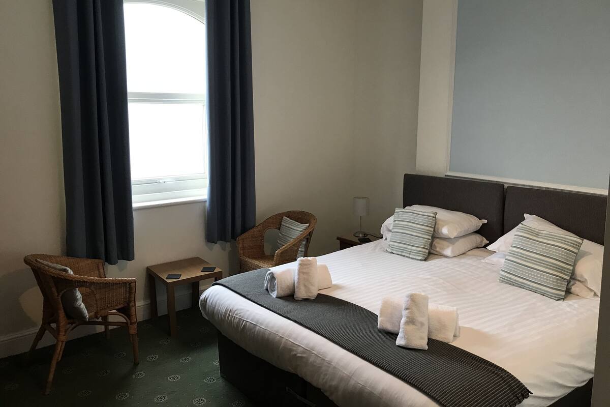 Quarto de hotel com uma cama, duas cadeiras, uma mesinha e uma janela com cortinas.