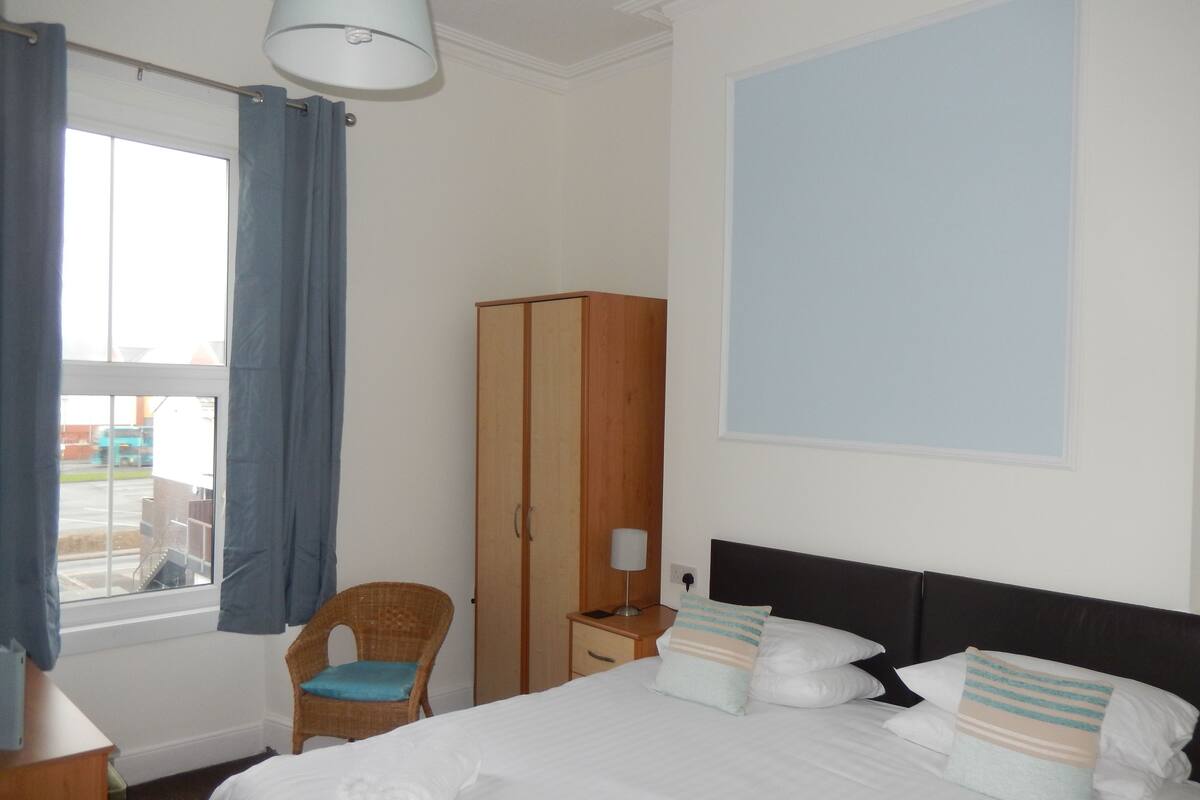 Quarto de hotel com cama, guarda-roupa, cadeira e janela com cortinas.