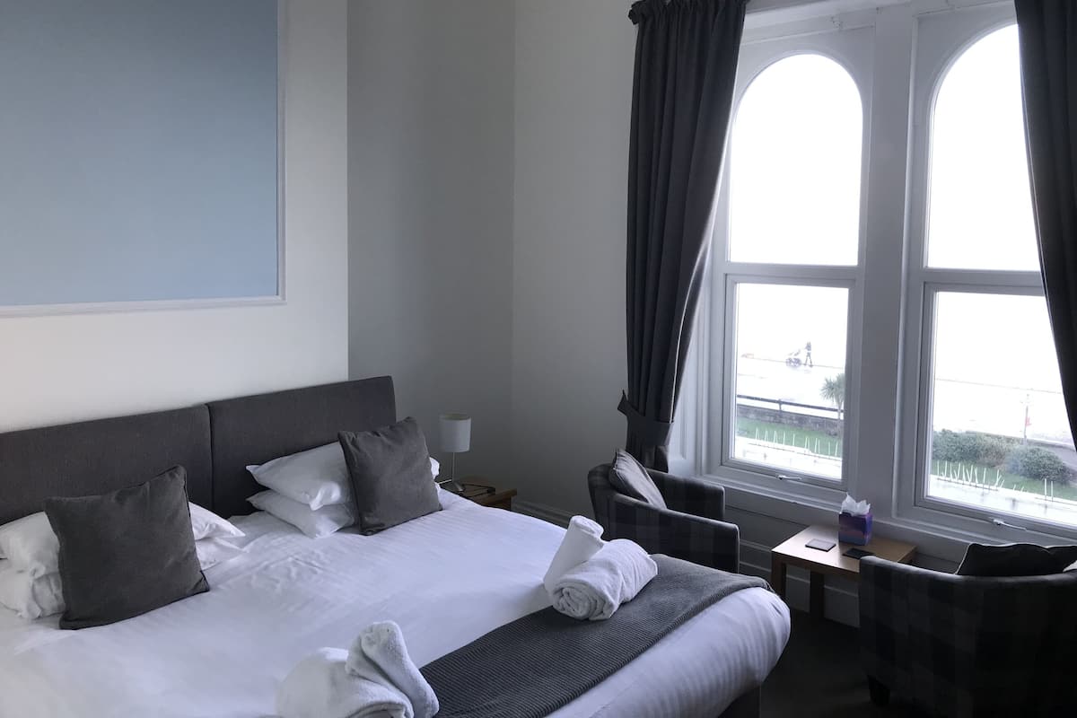 Quarto de hotel com cama, uma cadeira, escrivaninha e janela com cortinas.