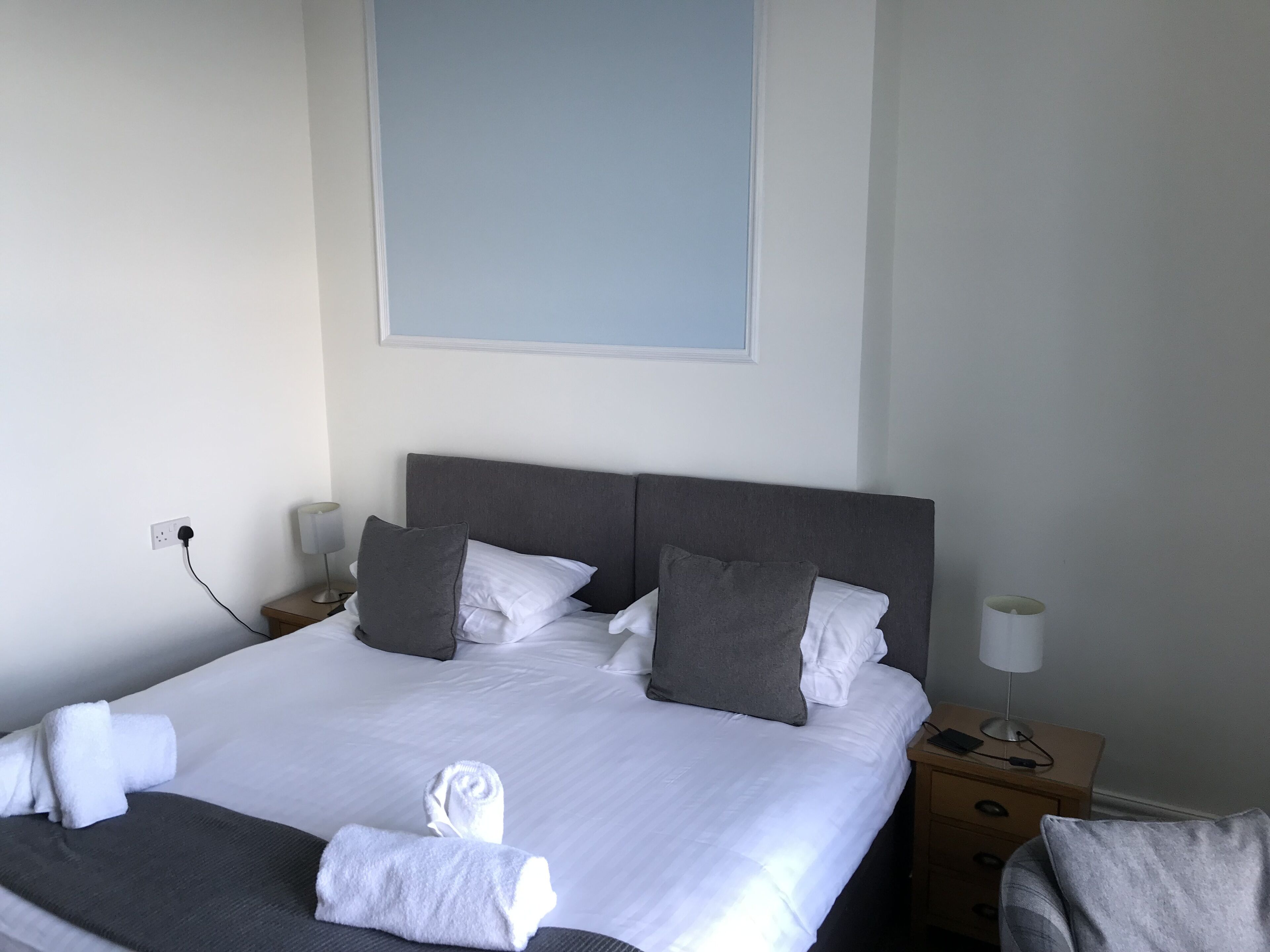 Een opgemaakt bed met witte en grijze lakens, een nachtkastje met een lamp en een stopcontact aan de muur.