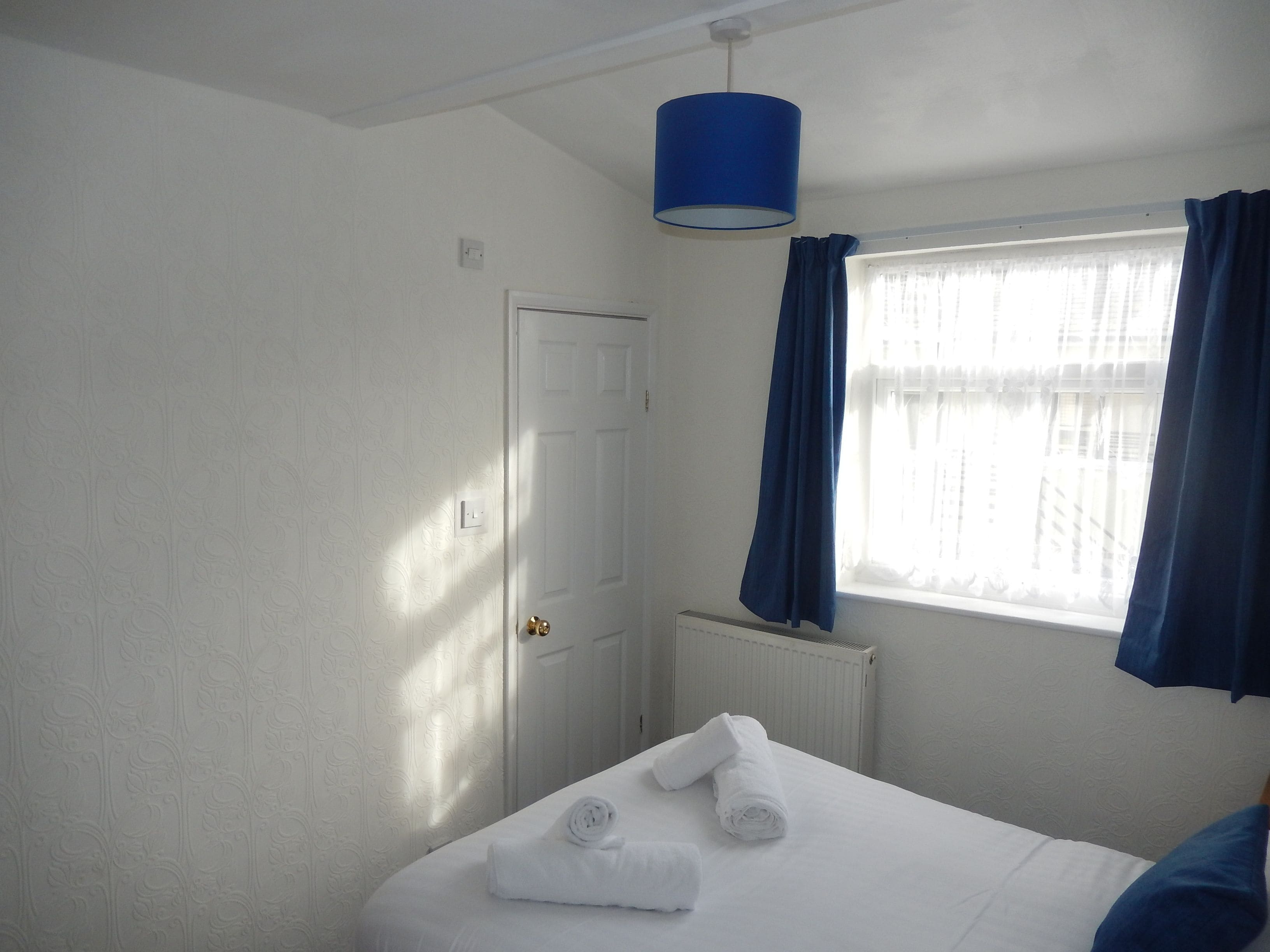 A hotel room with a bed, a white door, a window with blue curtains, and a blue hanging light fixture.