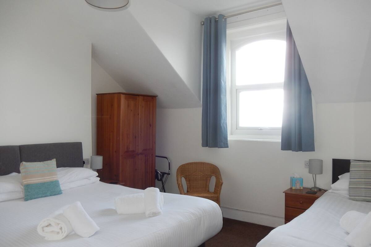 Quarto de hotel com duas camas, um guarda-roupa de madeira, uma cadeira de vime, uma mesa de cabeceira com abajur e uma janela com cortinas azuis.