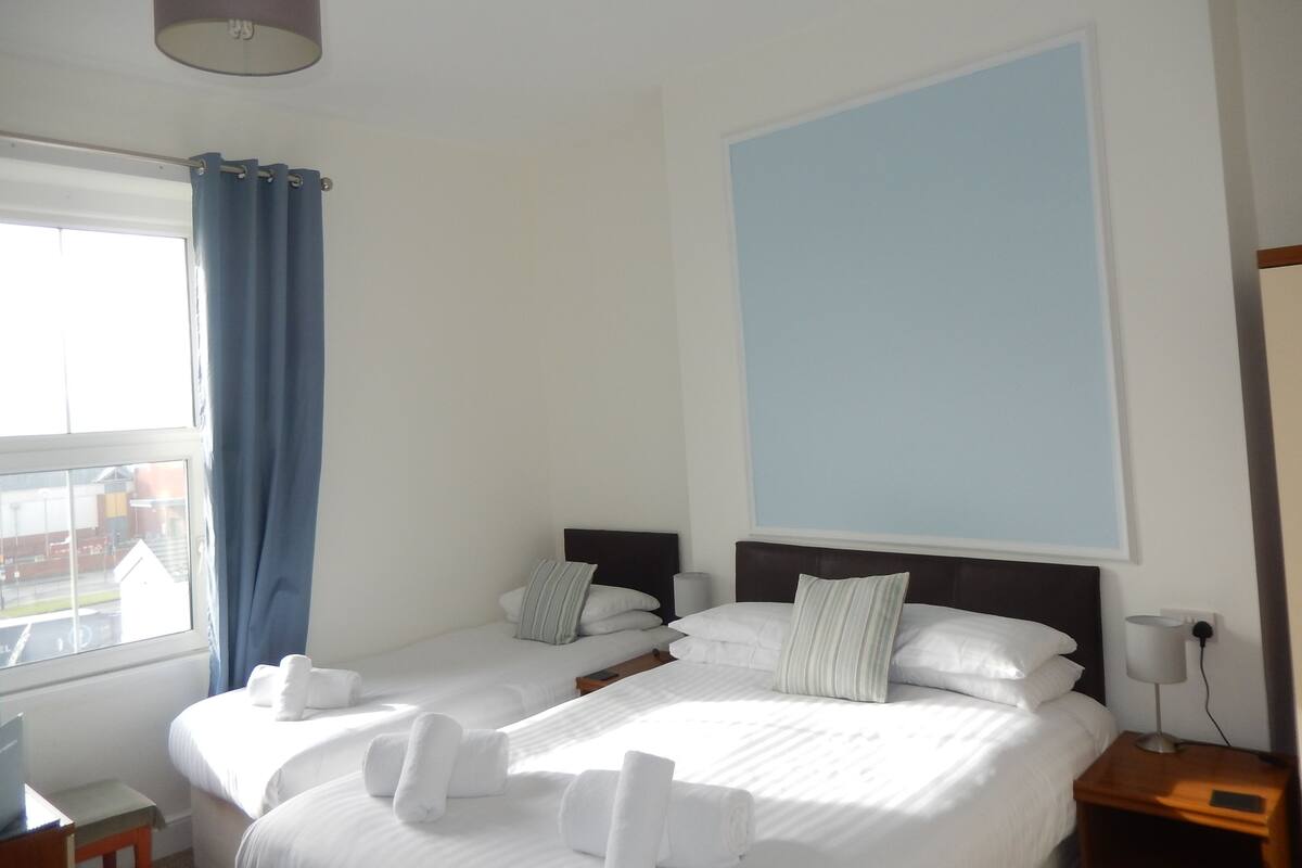 Quarto de hotel com duas camas, uma janela com cortinas azuis, uma luminária na mesinha de cabeceira e vista para o exterior.