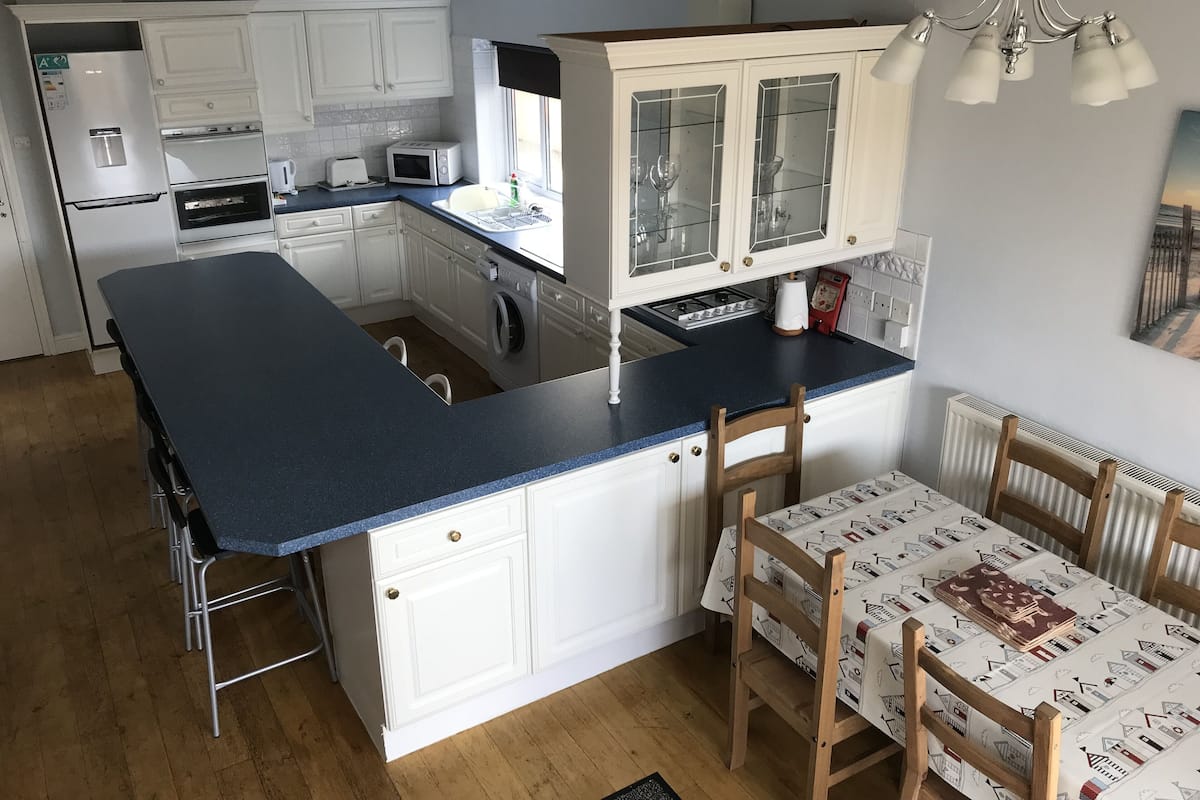 Uma cozinha compacta com armários brancos, bancada azul e um espaço de refeições com mesa coberta por uma toalha de mesa estampada com motivos domésticos.