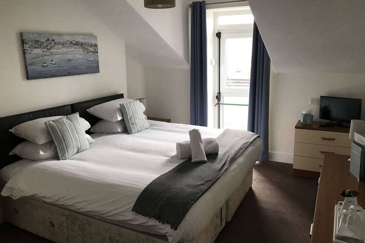 Cama bem arrumada com almofadas e um edredom, um quadro na parede, televisão sobre uma mesa de madeira e uma porta com cortinas.