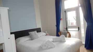 Habitación de hotel con cama, ropa de cama blanca, mesita de noche, ventana con cortinas azules y vistas al exterior.