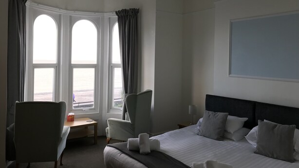Habitación de hotel con una cama grande, un escritorio, dos sillas y vista al mar a través de tres ventanas.