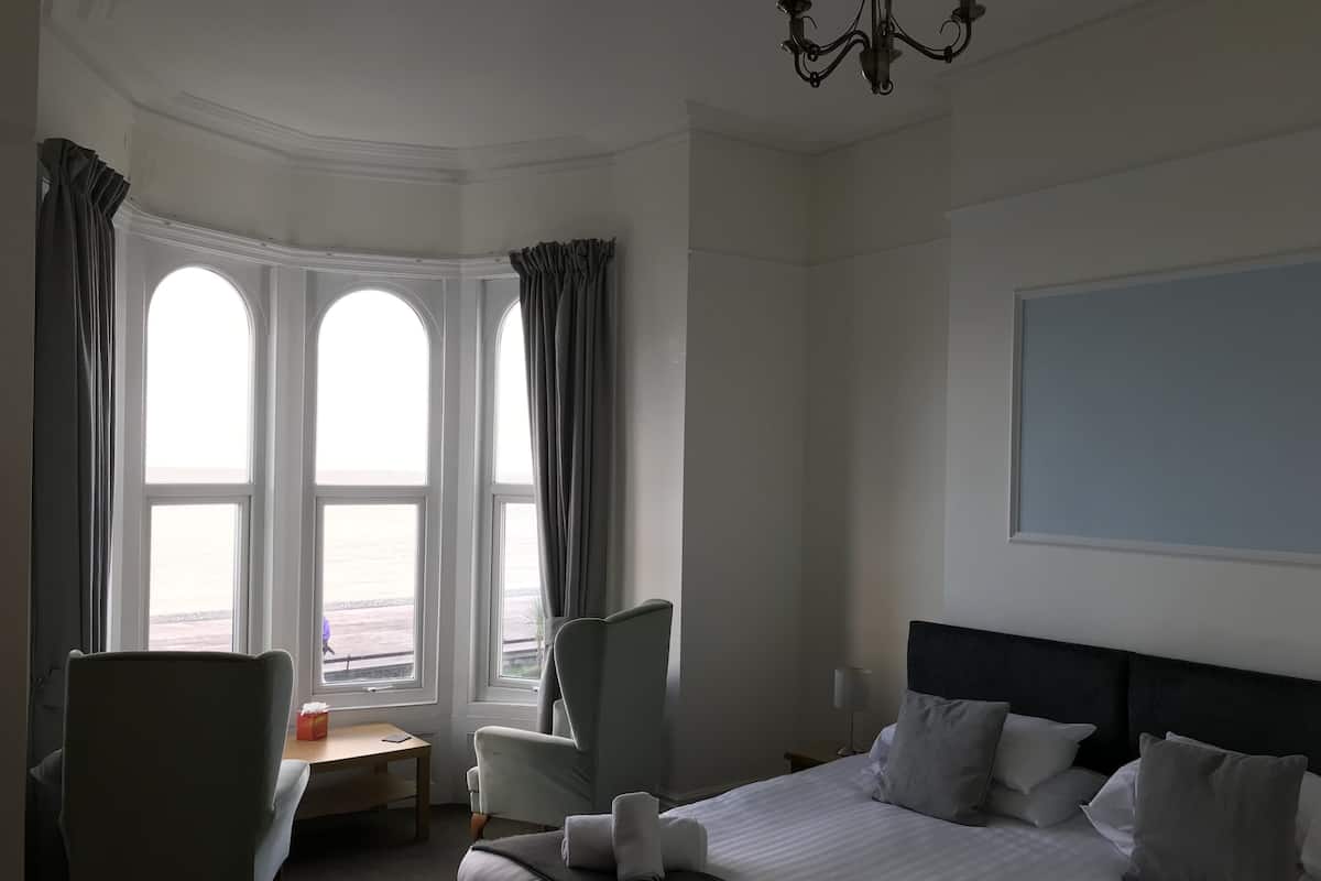 Quarto de hotel com uma cama grande, uma escrivaninha, duas cadeiras e vista para o mar através de três janelas.