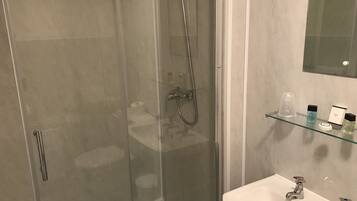Un baño moderno con ducha de vidrio, lavabo blanco con dos grifos y repisa de vidrio con artículos de aseo.