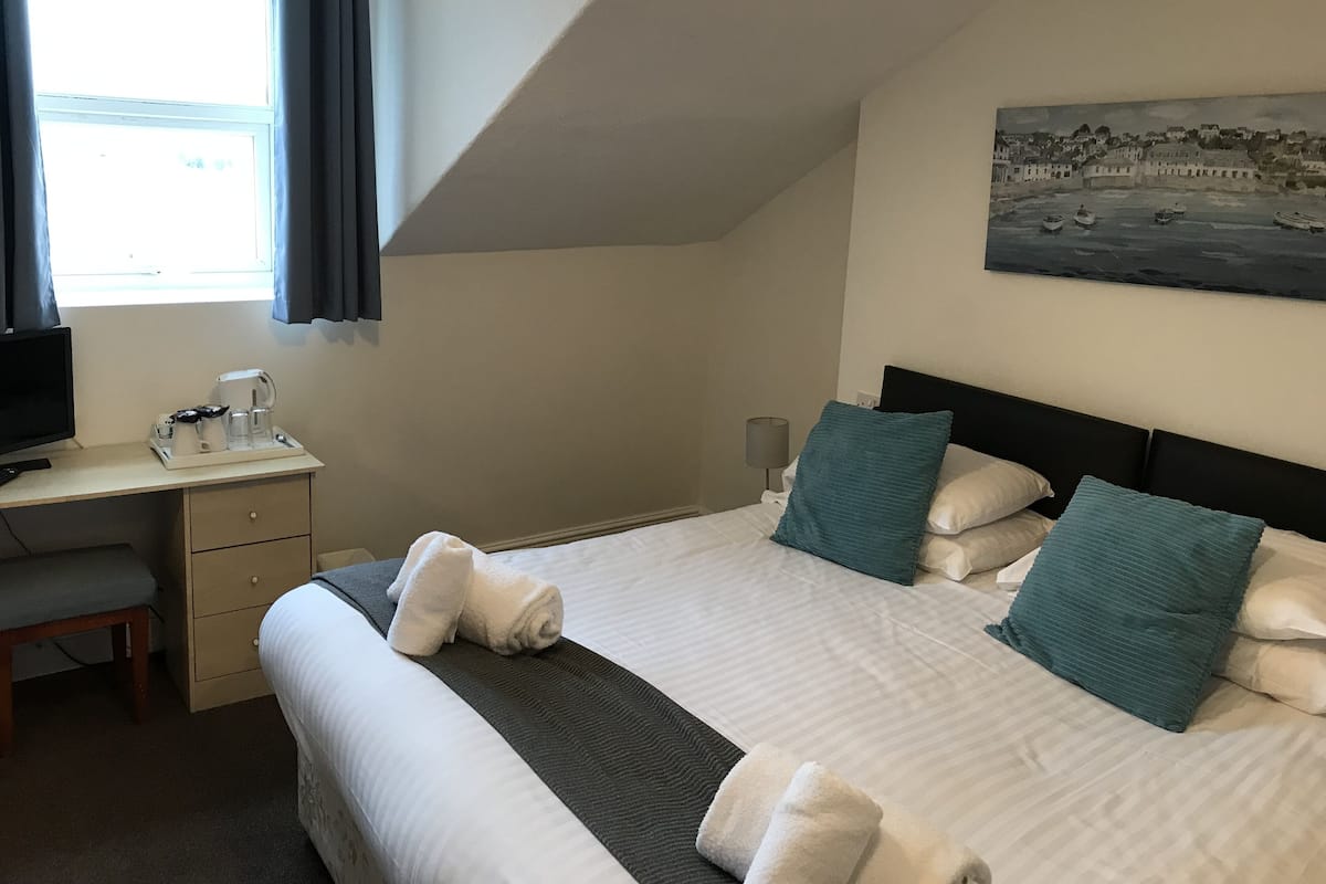 Quarto de hotel com uma cama, uma escrivaninha com computador, uma cadeira, um quadro na parede e uma janela com cortinas.