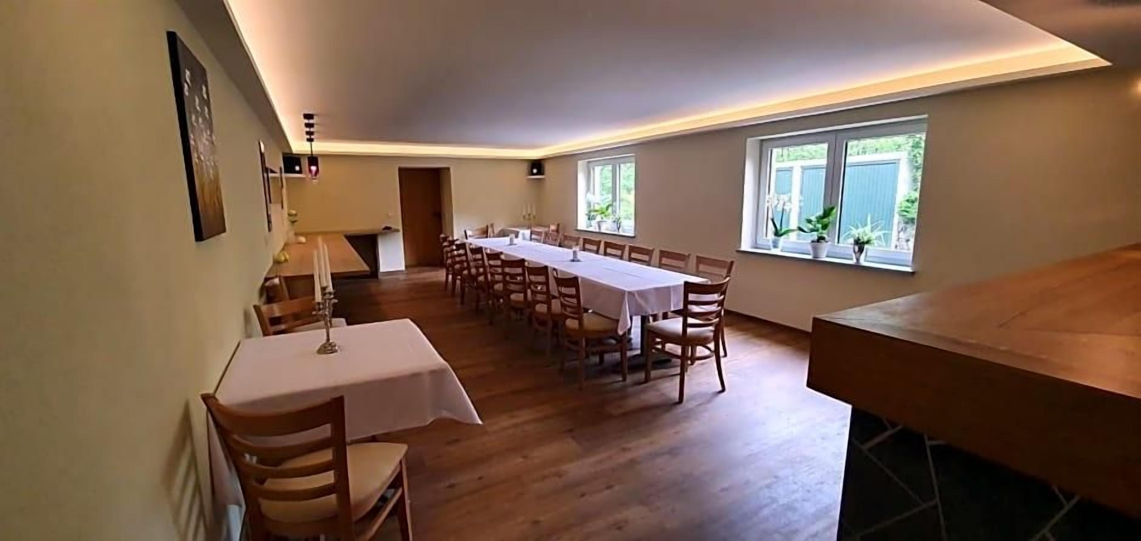 A dining area with a long table set for multiple guests, wooden flooring, and windows providing natural light.