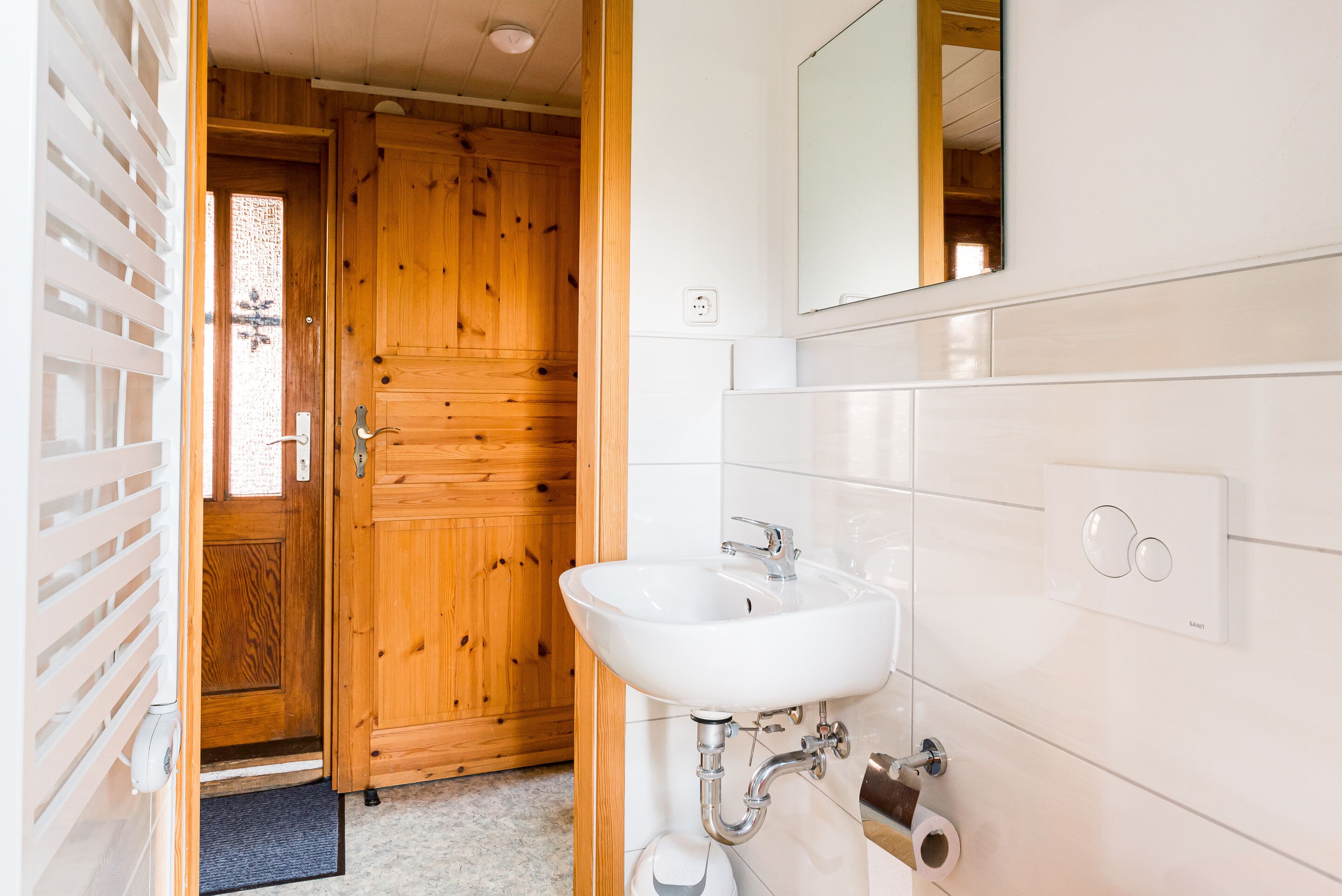 A bathroom with a wooden door, white tiles, a sink, and a mirror.