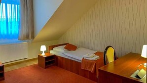 A small, triangular room with a single bed, a desk, and a chair. There is a window with curtains, a radiator, and a patterned wall.