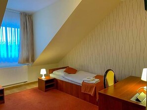 A small, triangular room with a single bed, a desk, and a chair. There is a window with curtains, a radiator, and a patterned wall.
