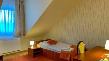 Ein kleiner, dreieckiger Raum mit einem Einzelbett, einem Schreibtisch und einem Stuhl. Es gibt ein Fenster mit Vorhängen, eine Heizung und eine Wand mit Muster.