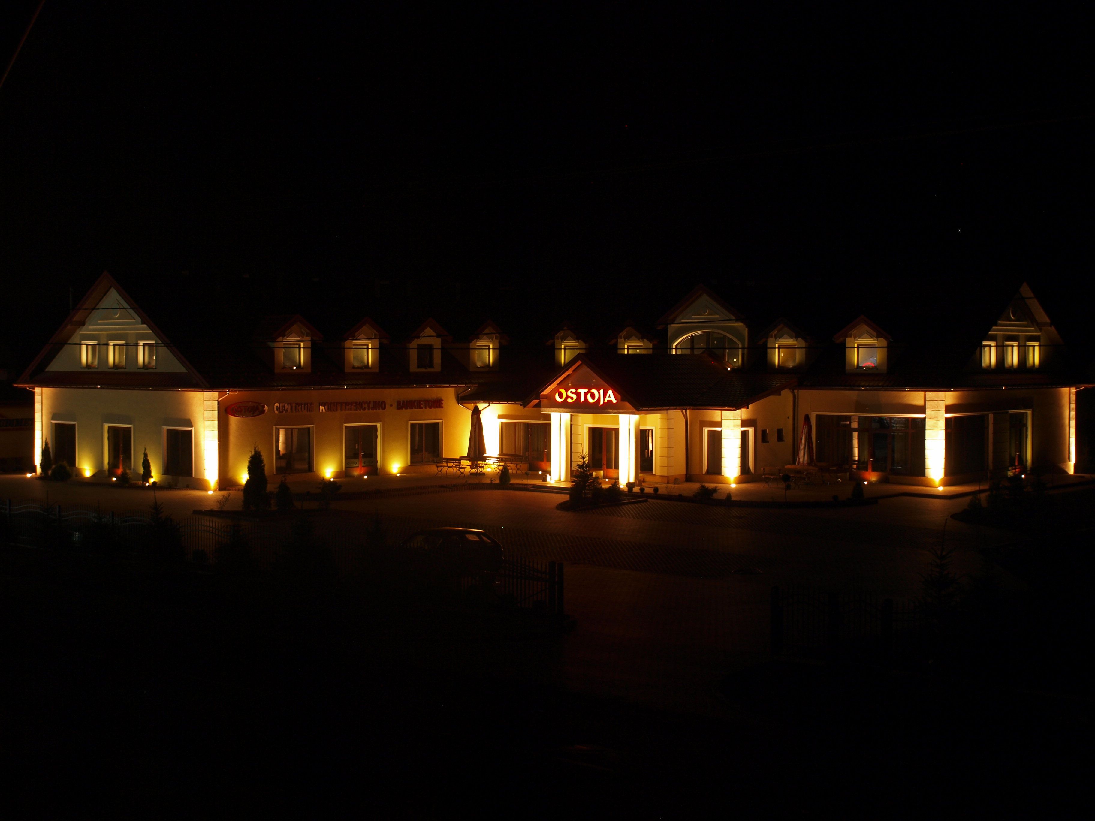 A hotel with a central building featuring a prominent 'OSTOJA' sign, multiple smaller attached structures, and a well-lit entrance area.