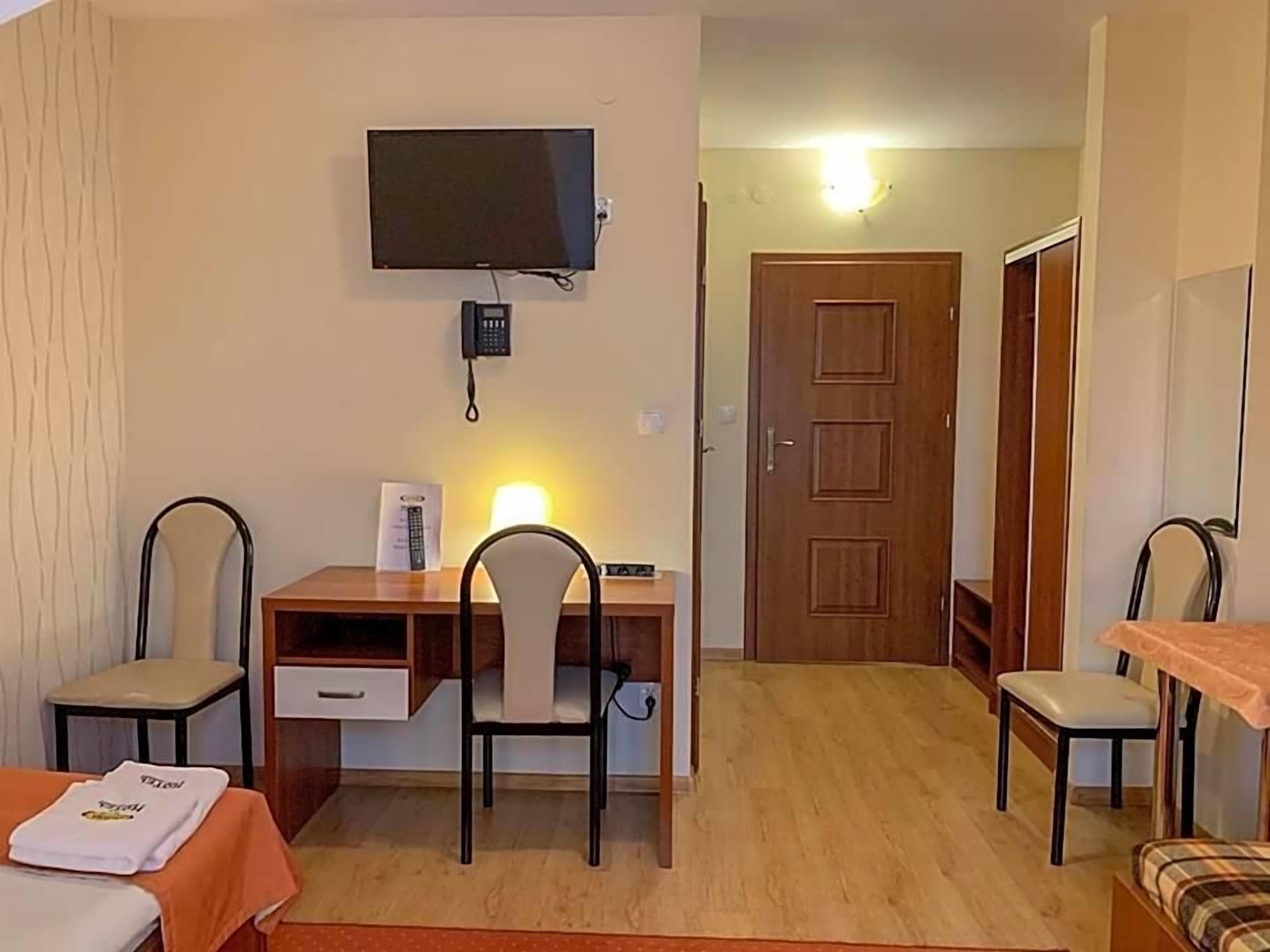 A hotel room with a bed, a desk with a chair, a television mounted on the wall, and a door leading to another room.