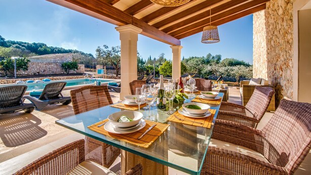 Una mesa de comedor con cubierta de vidrio, sillas de mimbre, una pared de piedra y vistas a una piscina y follaje.