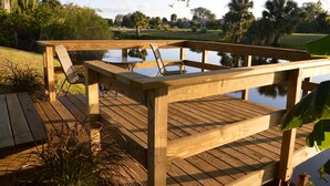 A wooden deck with a table and chairs, overlooking a calm body of water and a grassy area with trees.