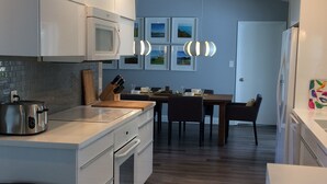 A modern kitchen with white cabinetry, a built-in oven, a microwave, and a stainless steel toaster. There is a dining area with a wooden table and chairs, and a wall-mounted light fixture.