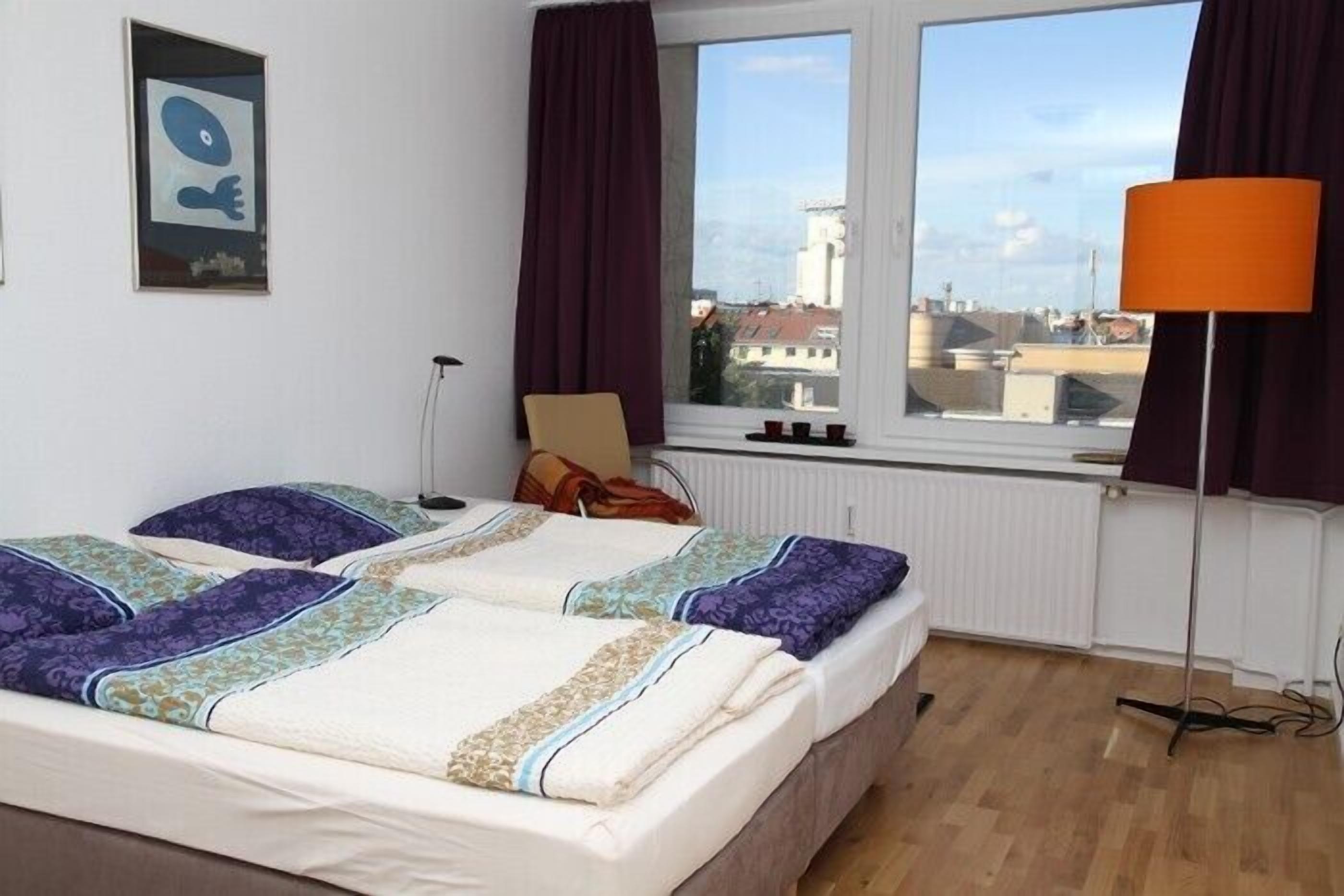 Ein Hotelzimmer mit zwei Betten, einem Fenster mit Blick auf Gebäude, einer Lampe und einem Bild an der Wand.