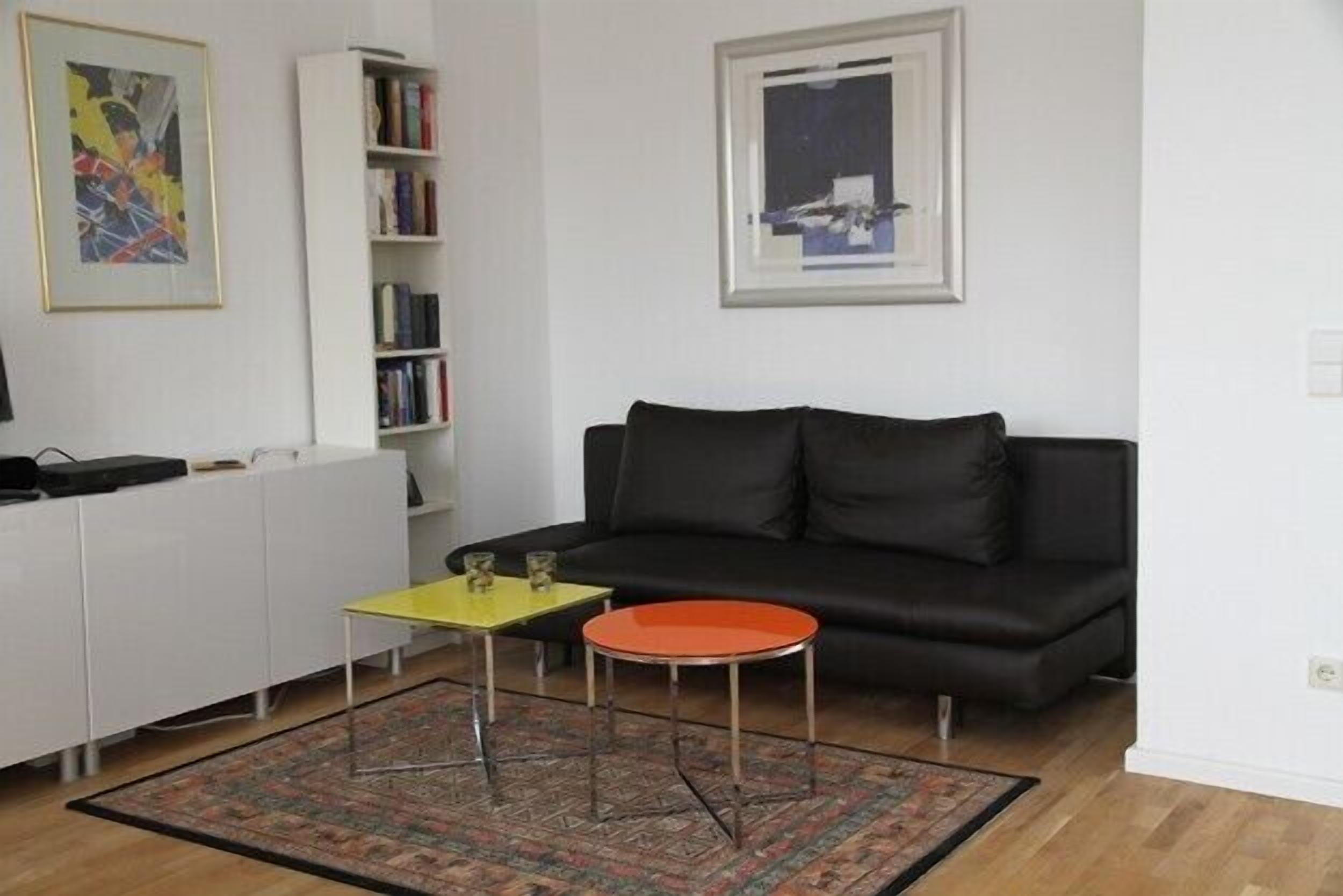 Ein Wohnzimmer mit einem schwarzen Sofa, einem gelben Couchtisch, einem roten Beistelltisch, einer weißen Kommode und einem Bücherregal.