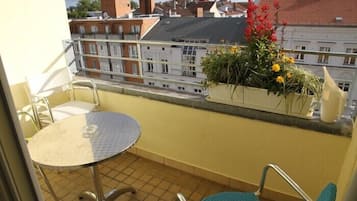 Ein Balkon mit rundem Tisch und zwei Stühlen, einem Blumentopf mit Blumen und Blick auf Gebäude im Hintergrund.