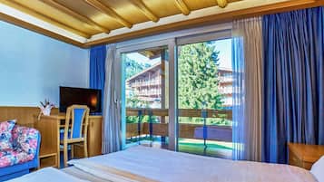 Une chambre d’hôtel avec un lit, un bureau et une chaise, une télévision, et une vue sur un immeuble et des arbres par la fenêtre.