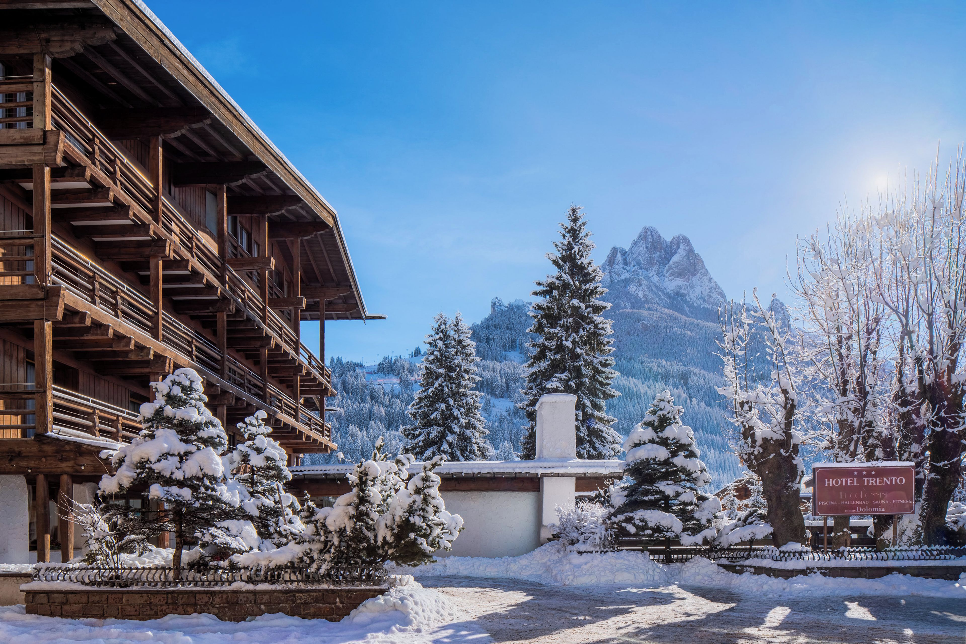 Ein Holzhaus mit einem Schild für Hotel Trento, umgeben von schneebedeckten Bäumen und Bergen im Hintergrund.