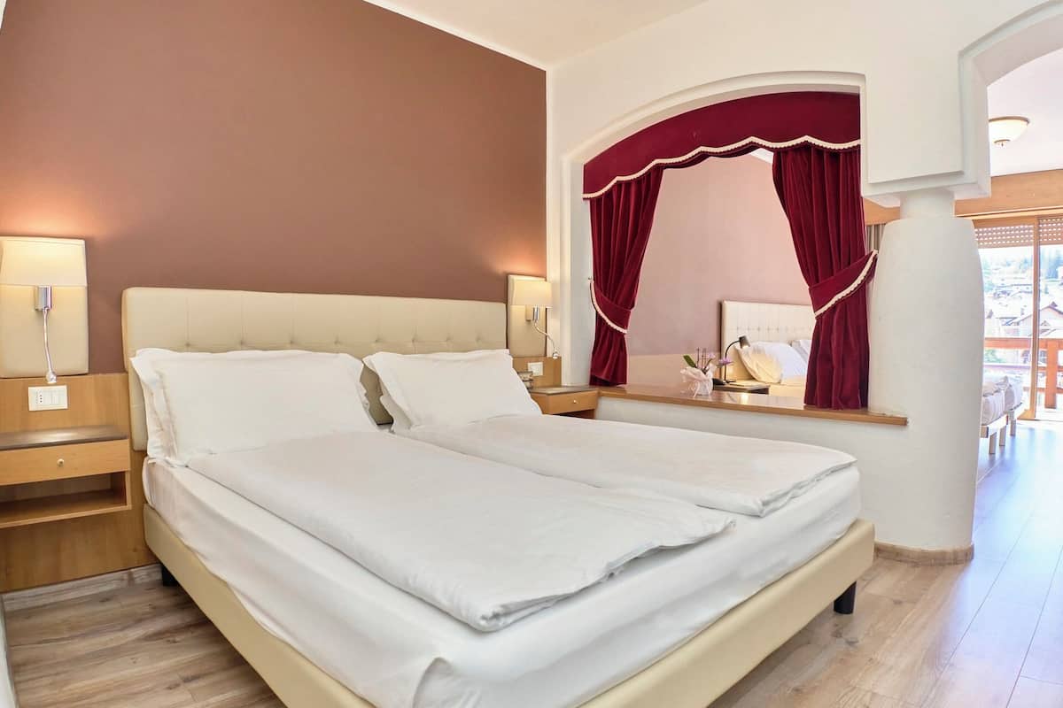 Un lit double avec du linge de lit blanc, une table de chevet avec une lampe et un miroir avec un rideau rouge.