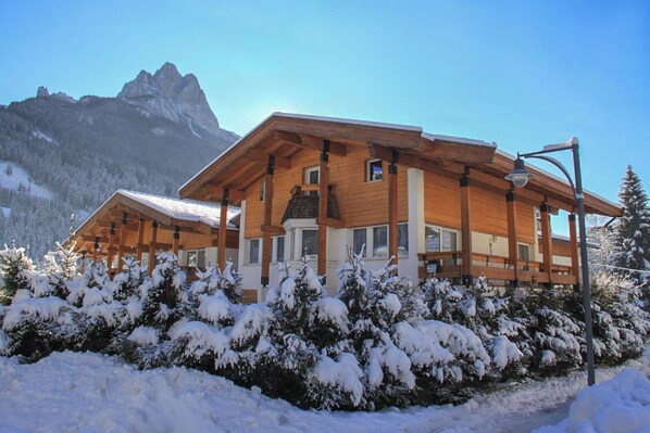 A wooden chalet with a sloped roof, surrounded by snow-covered trees and a mountain in the background.