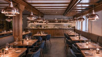 Un interior de restaurante con varias mesas para comer, sillas, iluminación colgante y una zona de bar.
