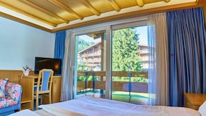 Hotelkamer met een bed, een televisie, een bureau en een stoel. Er is een schuifdeur die leidt naar een buitenruimte met uitzicht op een gebouw en bomen.