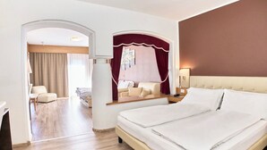 Een hotelkamer met een groot bed, een zithoek met een bank en fauteuils, een eettafel en een uitzicht door een boogpoort naar een balkon.