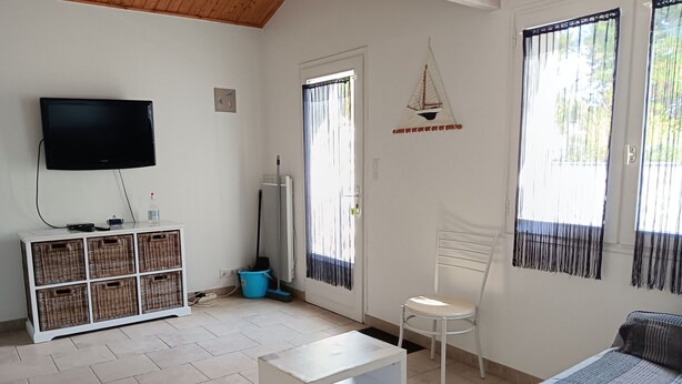 A room with a flat-screen TV, wicker storage unit, white chair, and a bed with a striped blanket. - Quiet cottage for 4 in La Faute-sur-Mer (La Faute-sur-Mer)