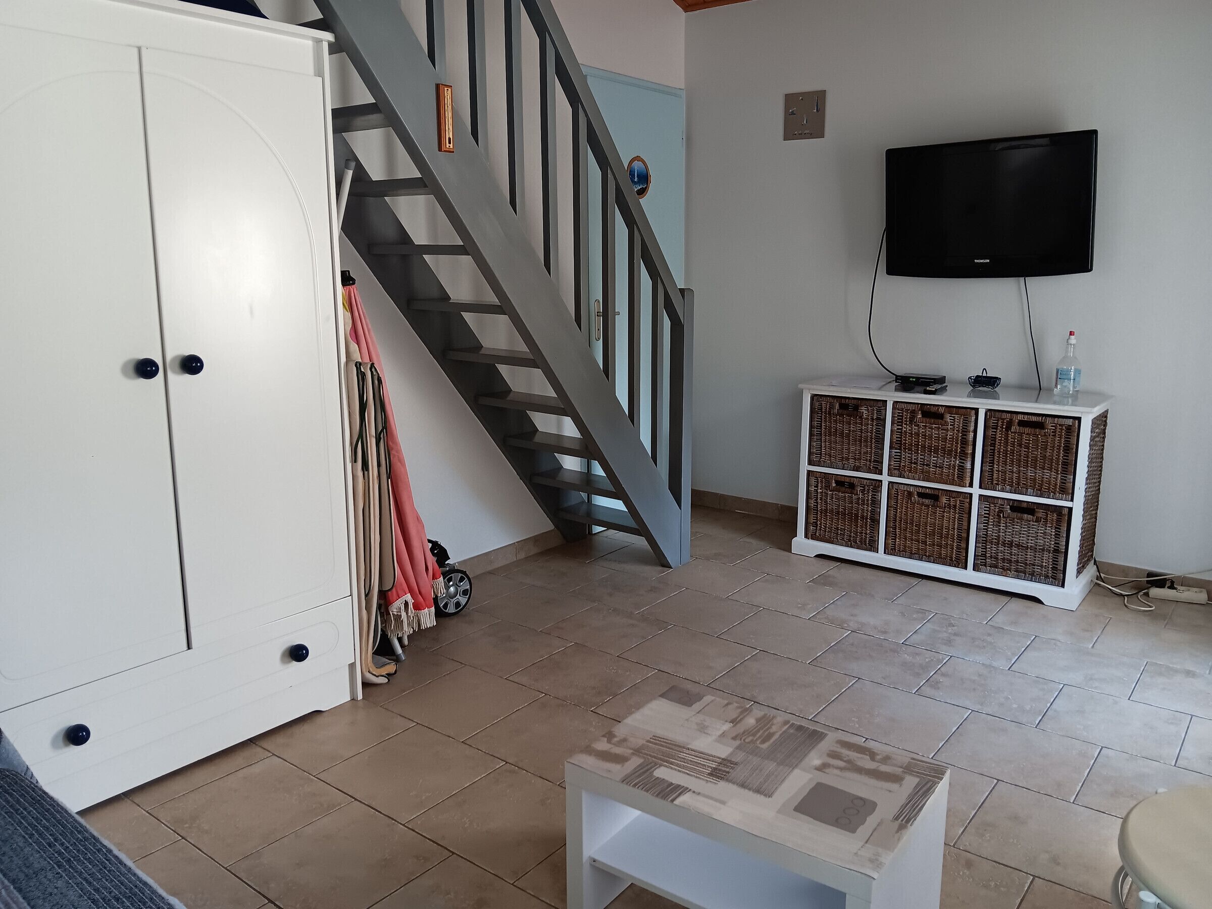 Un salon avec un escalier, un téléviseur fixé au mur et un meuble blanc contenant des paniers en osier.