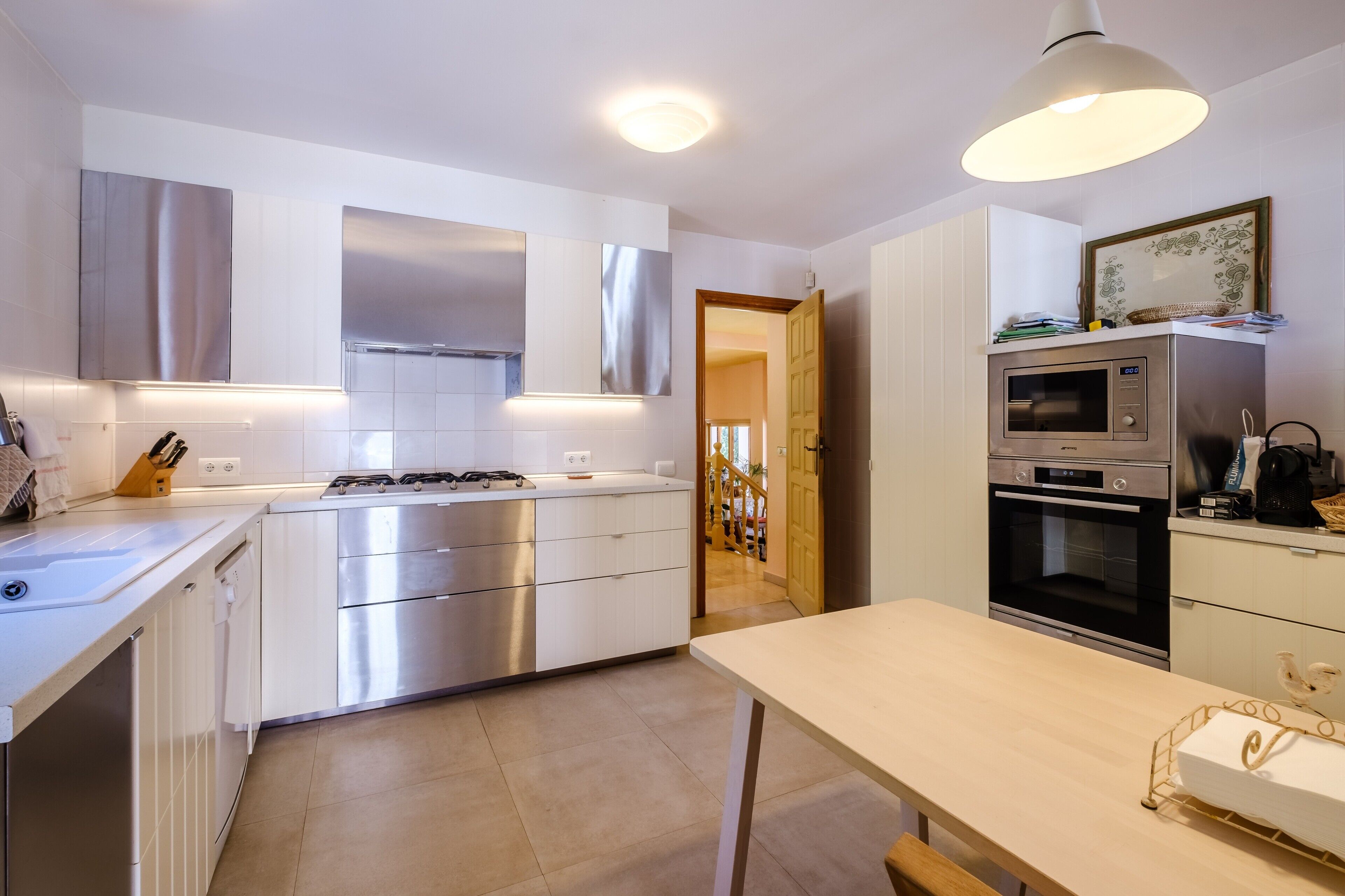 Een moderne keuken met roestvrijstalen apparatuur, een eethoek met tafel, en een zicht op een gang.