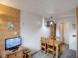 A room with a wooden TV stand, a small table, and a dining table with chairs. There is a wall clock and a wooden cabinet.