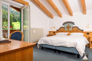 A bedroom with a bed, bedside tables, a wooden desk, and a view of a forest through a window. - Dolomites Hotel Valacia  (San Giovanni di Fassa)