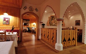 A restaurant interior with wooden walls, a central archway, and a dining area with tables and chairs. - Dolomites Hotel Valacia  (San Giovanni di Fassa)