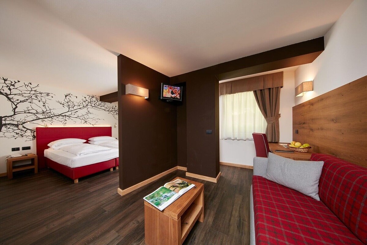 A hotel room with a red bed, a wooden headboard, a wooden coffee table with a magazine, a red sofa, and a wall-mounted TV.