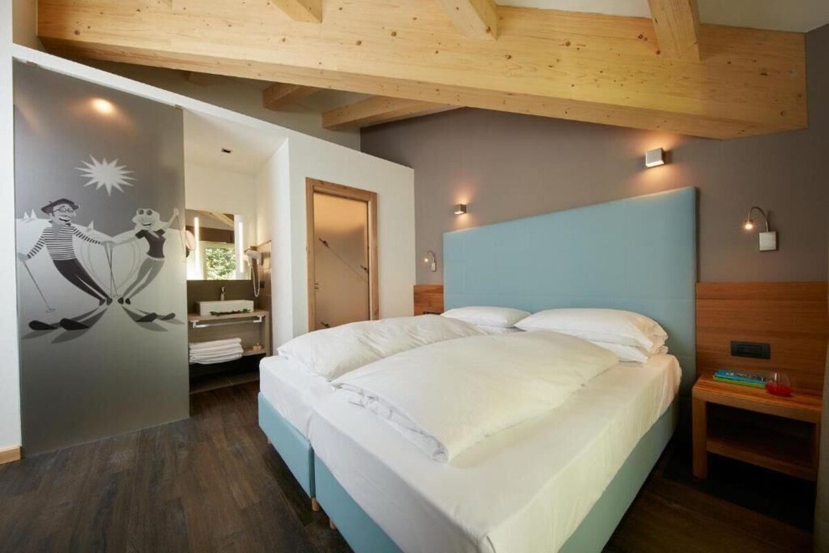 A bedroom with a large bed, wooden ceiling beams, a wall-mounted light, and a bathroom with a mirror and sink.