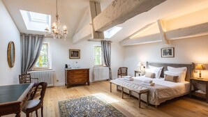 Une chambre à coucher avec un grand lit, une commode en bois, un bureau et une chaise. Il y a deux fenêtres avec des rideaux, un lustre et un radiateur.